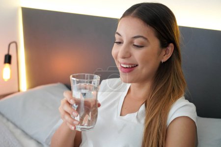 Foto de Una joven en la cama bebe un vaso de agua que acaba de despertarse por la mañana. Importancia de una buena hidratación diaria. - Imagen libre de derechos
