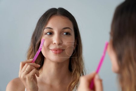 Photo for Facial hair removal. Dermaplaning. Close-up of beautiful teenager girl shaving her face by razor at home. Pretty young woman using razor on bathroom. - Royalty Free Image
