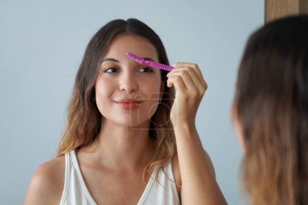 Photo for Dermaplaning. Pretty hispanic girl shaving her eyebrows by razor on the mirror at home. Facial hair removal. - Royalty Free Image
