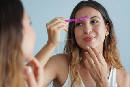 Photo for Dermaplaning. Beautiful young woman shaving her eyebrows by razor on the mirror at home. Facial hair removal. - Royalty Free Image