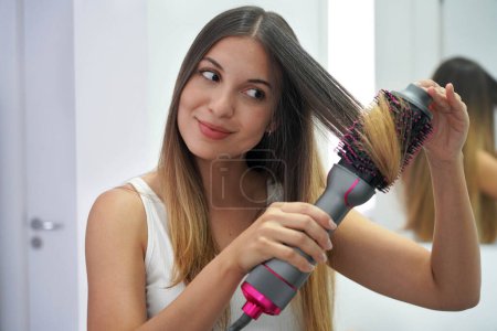 Hot air hair brush. Close-up of young woman using round brush hair dryer to style hair. Pretty girl using electric blowout brush hair dryer.