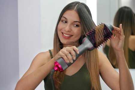 Porträt einer jungen Frau, die einen runden Bürstenhaartrockner benutzt, um die Haare zu Hause auf einfache Weise zu stylen. Mädchen mit elektrischer Ausblasbürste Haartrockner. Heißluft-Haarbürstenkonzept.