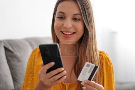 Gros plan d'une jeune femme souriante tenant sa carte de crédit et regardant son smartphone à la maison