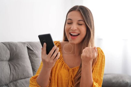 Excited young woman with eyes closed and fist up holds smartphone on sofa at home