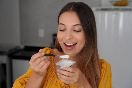 Atractiva joven sonriente comiendo yogur natural en la cocina en casa