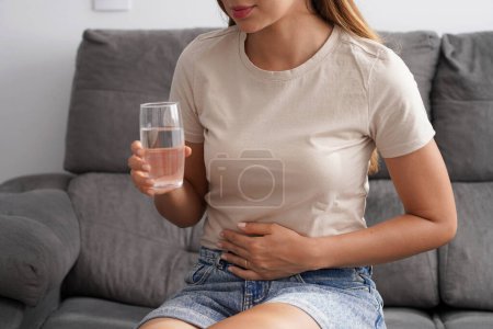 Costiveness. Mujer joven irreconizable que sufre de dolor abdominal sostiene un vaso de agua en su mano en casa.