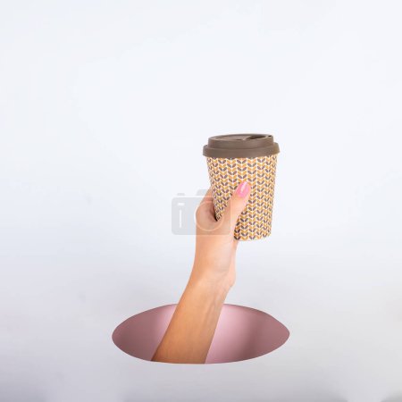 Téléchargez les photos : Main de femme à l'extérieur d'un trou tenant une tasse de café sur fond rose et bleu. Photo de haute qualité - en image libre de droit