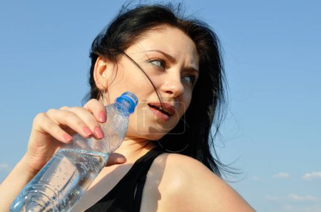 Photo for Young attractive woman drinks cold water in hot day on blue sky background - Royalty Free Image