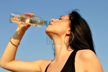 Photo for Young attractive woman drinks cold water in hot day on blue sky background - Royalty Free Image