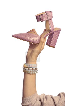 Photo for A female hand with a set of bracelets holding a pink high-heeled pointed-toe crocodile leather summer shoe isolated on a white background. Graceful hand showcasing footwear - Royalty Free Image