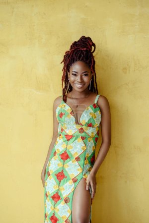 Foto de La atractiva joven negra africana vistiendo un adorno multicolor vestido de raja alta, mirando a la cámara y sonriendo, aislada sobre un fondo de pared amarillo con espacio para copiar. Bastante modelo étnico - Imagen libre de derechos