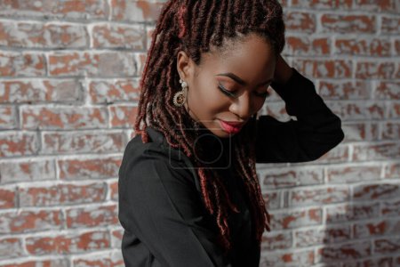 A portrait of pretty african woman with flawless skin, dreadlocks hairdo and stylish makeup, wearing black shirt and eloquent earrings, looking down against a brick wall with copy space.