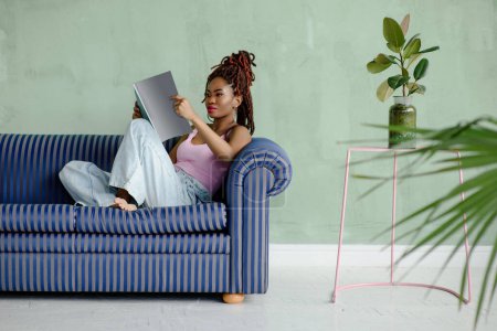 Foto de Mujer joven bastante africana leyendo revista y sonriendo mientras se sienta en el sofá en una cómoda pose en una sala de estar con paredes de color verde claro y plantas de interior. Concepto de ocio. - Imagen libre de derechos