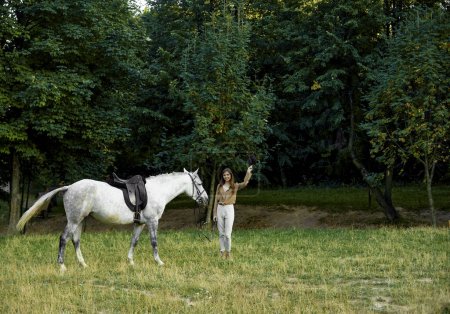Téléchargez les photos : Une jolie fille debout avec un cheval gris sellé sur un champ d'herbe avec des arbres derrière elle et enlevant son chapeau, faisant un salut. Passer un week-end dans la nature avec un cheval de compagnie. Thérapie du cheval - en image libre de droit