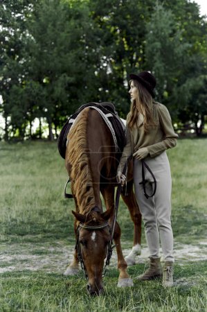 Téléchargez les photos : Jolie femme au chapeau noir avec un cheval brun paît un cheval à la campagne. Les sports de montagne contribuent à une bonne santé et à la relaxation. Certificat cadeau pour émotions, hippothérapie - en image libre de droit
