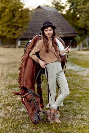 Téléchargez les photos : Jeune femme au chapeau noir se tient sur l'herbe près d'un cheval brun pâturage herbe. Des chevaux nourris. Certificat cadeau pour l'hippothérapie et les émotions. Cheval au coucher du soleil le soir - en image libre de droit