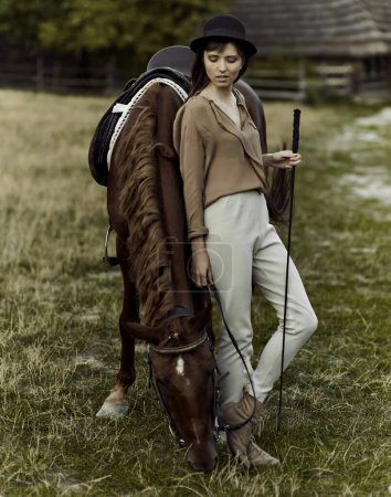 Téléchargez les photos : Une jeune femme au chapeau noir paît un cheval brun. Une femme et son cheval à la campagne, sur un champ herbeux. - en image libre de droit