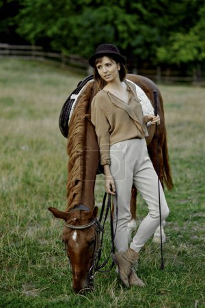 Téléchargez les photos : Femme à la haine noire se tient près de pâturage cheval brun sur l'herbe. Femme se lie avec son cheval sur un terrain herbeux. - en image libre de droit