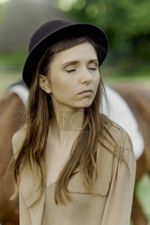 Téléchargez les photos : Portrait d'une jeune cavalière calme aux yeux fermés avec un cheval en arrière-plan. Prise de vue d'une jolie jeune femme debout devant son cheval à l'extérieur. Certificat-cadeau aventure équestre - en image libre de droit