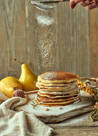 Photo for Powdered sugar is sprinkled on a pile of buttermilk pancakes with a pair of pears in the distance a small branch of sea buckthorn, and a spoon for drizzling honey. Lifestyle food photography. - Royalty Free Image