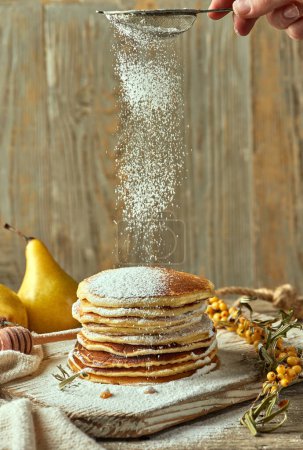 Photo for Vanilla pancakes on a light-colored wooden table surrounded by a pair of pears in the distance, a small sea buckthorn branch, and a spoon for pouring honey. Powdered sugar is sprinkled. Food blog. - Royalty Free Image