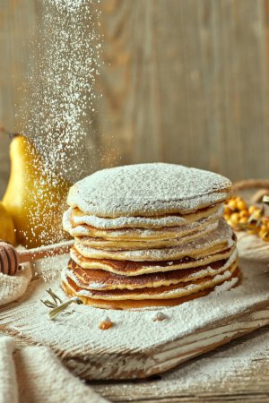 Photo for Closeup shot of homemade oatmeal pancakes dusted with powdered sugar stacked on a pale wooden platter. Pears and sea buckthorn berries in the backdrop. An element of festivity. Food photography. - Royalty Free Image