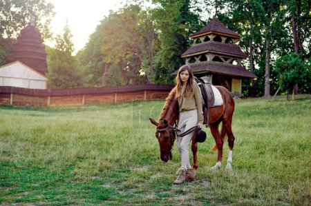 Téléchargez les photos : Une jeune femme avec un cheval à la campagne sur un terrain herbeux. Jeune jockey dressant des animaux, une ferme de cavaliers et d'athlètes, et une arène de plein air simulée pour les sports de selle avec des chevaux - en image libre de droit
