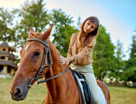 Téléchargez les photos : Une femme caressant doucement un cheval sur un champ herbeux au milieu des paysages ruraux. Un portrait d'un jeune jockey met en valeur la formation à la ferme et une installation sportive de selle en plein air. Équitation, équitation - en image libre de droit
