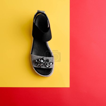 Foto de Elegante sandalia negro-plateada decorada con una dispersión de pedrería sobre fondo amarillo y rojo. Blog de moda. venta de zapatos campaña de marketing venta al por menor de calzado, ortopedia, tienda de outlet de comercio electrónico - Imagen libre de derechos