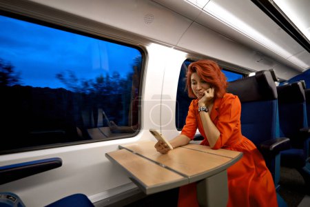 Foto de Feliz joven caucásica mujer pasajero sonríe utilizando el teléfono móvil inteligente en vagón de tren en su asiento azul. Disfrutando del concepto de viaje. 5g tecnología móvil en línea en viaje de transporte. - Imagen libre de derechos