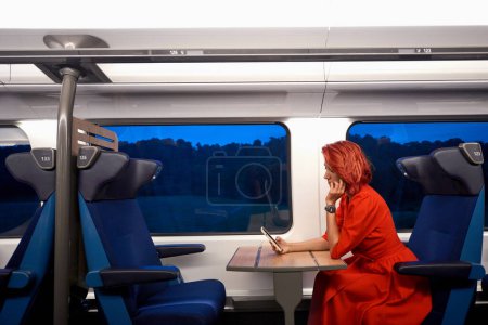 Foto de Vista lateral de calma pacífica impresionante hembra en buen equipo con teléfono inteligente que descansa en un cómodo sillón cerca de la ventana del tren. Distancia social para la prevención de enfermedades en el viaje público en tren. - Imagen libre de derechos
