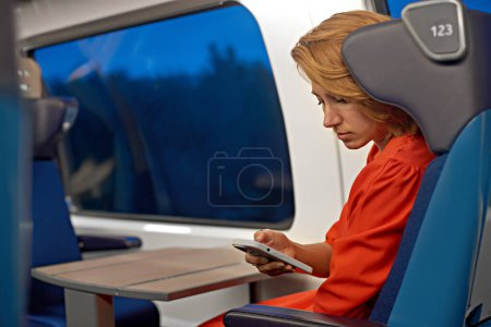 Foto de Una joven atractiva con el pelo rojo y un vestido rojo que trabaja en línea de forma remota desde su teléfono y viaja. Capacitar a los viajes y la lectura en las redes sociales de telefonía móvil, notificaciones, mensajes o vídeo. - Imagen libre de derechos