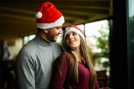 Photo for Beautiful couple hugging wearing Santa hats near a window - Royalty Free Image