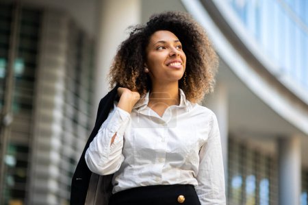 Téléchargez les photos : Confiante jeune afro-américaine gestionnaire en plein air dans un cadre urbain moderne tenant sa veste - en image libre de droit