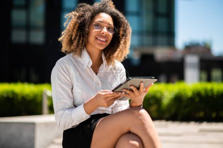 Foto de Mujer de negocios afroamericana sonriente usando una tableta digital al aire libre sentada en un banco - Imagen libre de derechos