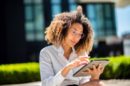 Téléchargez les photos : Femme d'affaires afro-américaine souriante utilisant une tablette numérique en plein air assise sur un banc - en image libre de droit