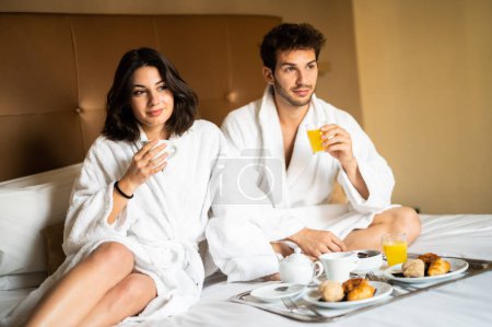Foto de Hermosa pareja desayunar en la cama en el hotel, hombre joven y mujer en albornoces felices juntos por la mañana - Imagen libre de derechos
