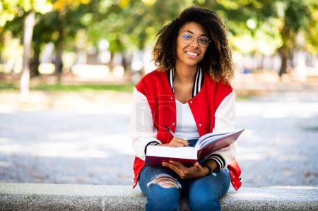 Téléchargez les photos : Belle étudiante africaine lisant un livre sur un banc dans un parc - en image libre de droit