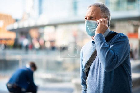 Téléchargez les photos : Homme masqué senior parlant au téléphone dans une place de la ville - en image libre de droit