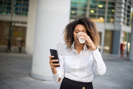 Téléchargez les photos : Portrait d'une femme d'affaires afro-américaine utilisant son téléphone portable dans un environnement professionnel tout en buvant du café - en image libre de droit