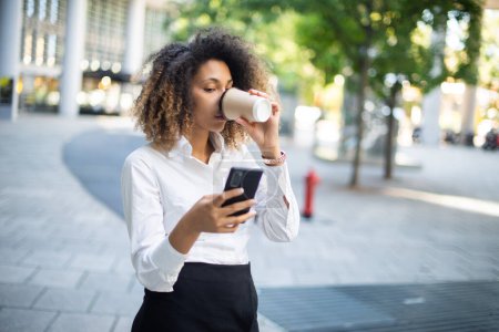 Téléchargez les photos : Portrait d'une femme d'affaires afro-américaine utilisant son téléphone portable dans un environnement professionnel tout en buvant du café - en image libre de droit