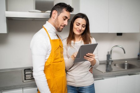Téléchargez les photos : Couple heureux à la recherche d'une recette sur la tablette pour cuisiner dans leur cuisine - en image libre de droit