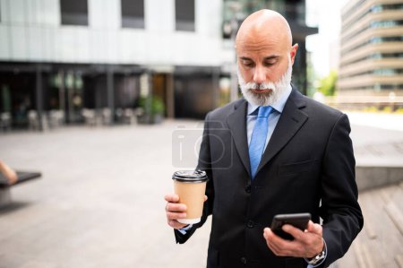 Foto de Elegante gerente calvo senior con barba blanca usando su teléfono inteligente al aire libre mientras bebe un café - Imagen libre de derechos