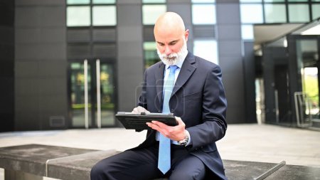 Téléchargez les photos : Gestionnaire élégant en utilisant sa tablette assise sur un banc en plein air dans une ville moderne - en image libre de droit