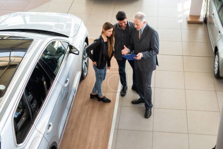 Téléchargez les photos : Joyeux jeune famille parlant au vendeur et choisissant leur nouvelle voiture dans un showroom - en image libre de droit