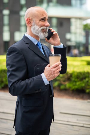 Photo for Bald businessman with a white beard talking on the phone and drinking coffee - Royalty Free Image