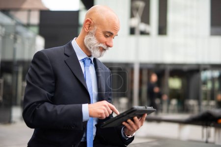 Foto de Hombre de negocios calvo con barba blanca usando su tableta en una ciudad moderna - Imagen libre de derechos