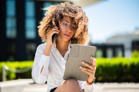 Foto de Sonriente mujer de negocios afroamericana en el teléfono usando una tableta digital al aire libre sentada en un banco - Imagen libre de derechos