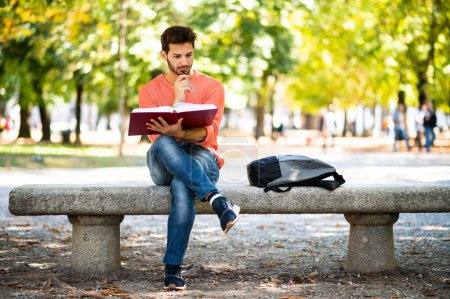 Photo for Handsome young man reading book on bench in the park - Royalty Free Image