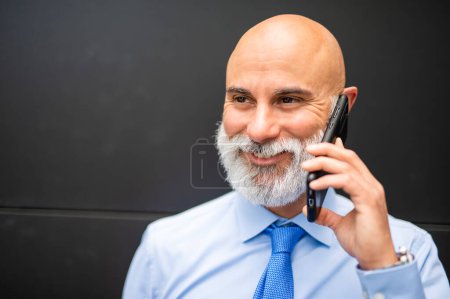 Foto de Calvo barbudo hombre de negocios de pie junto a una pared negra mientras habla por teléfono - Imagen libre de derechos